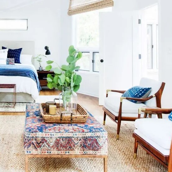 a coastal bedroom with a bed and printed bedding, white chairs, a boho printed ottoman, a jute rug and potted greenery