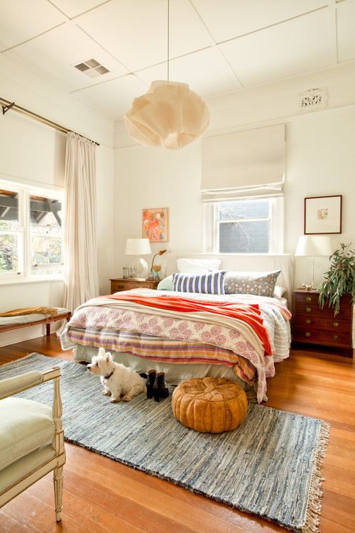 a cozy eclectic bedroom with a bed and bright bedding, stained nightstands, artwork, greenery and a chair