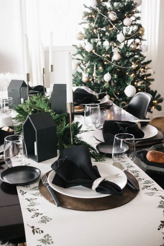 a modern black and white Christmas tablescape with a printed runner, black napkins, black house-shaped candleholders and evergreens and white candles