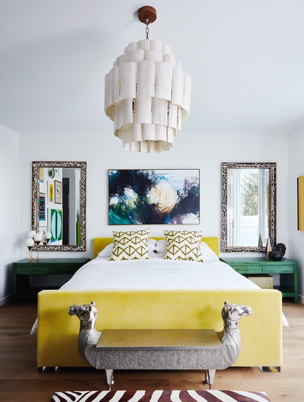 a stylish maximalist bedroom done in white, with a yellow upholstered bed, green nightstands, mirrors, a statement artwork and a catchy chandelier