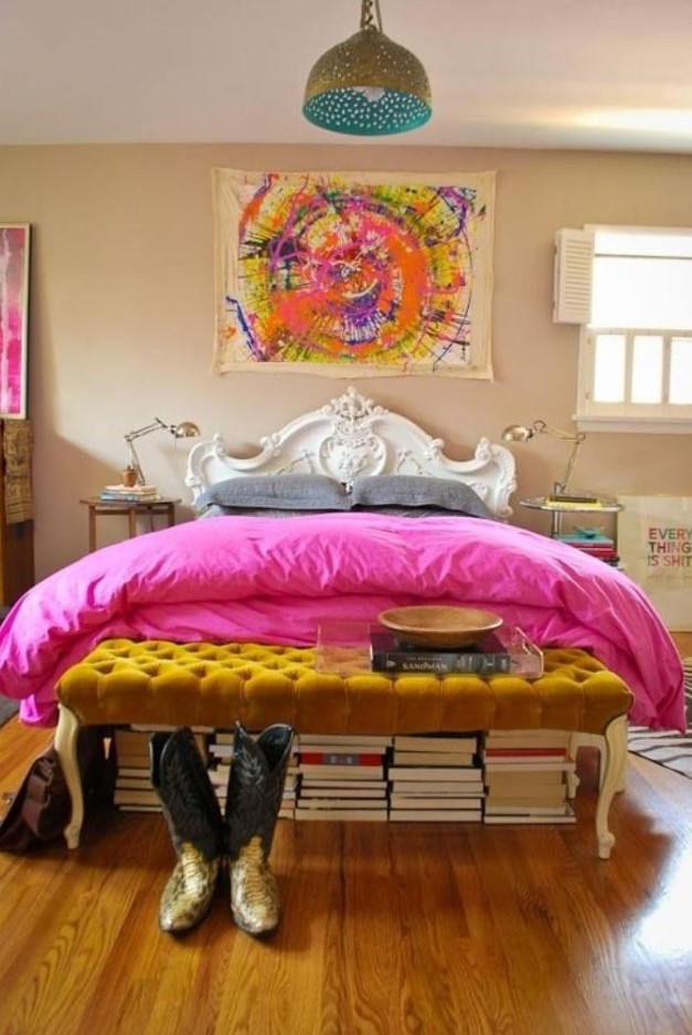 a super bright eclectic bedroom with bold artworks, a mustard tufted ottoman, stacks of books, a refined bed and mismatching nightstands