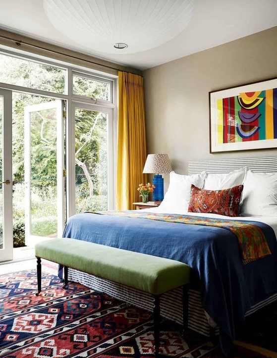 an eclectic and colorful bedroom with tan walls, a bed with colorful bedding, a green bench, a colorful boho rug and yellow curtains