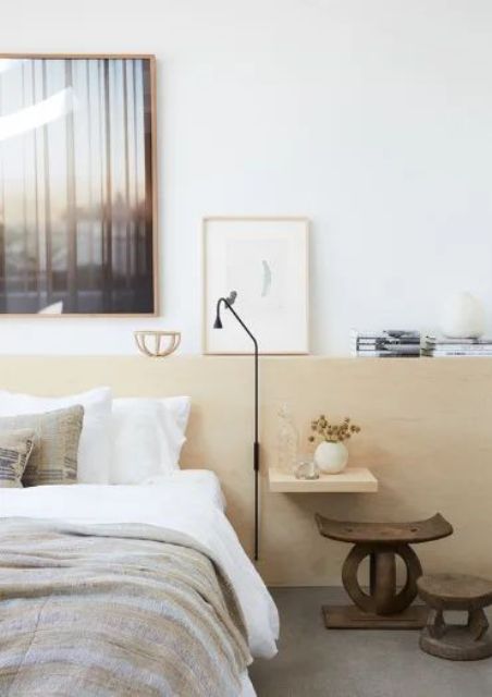 an eclectic bedroom with a bed and neutral bedding, a stained headboard panel for storage, dark-stained stools and a sconce