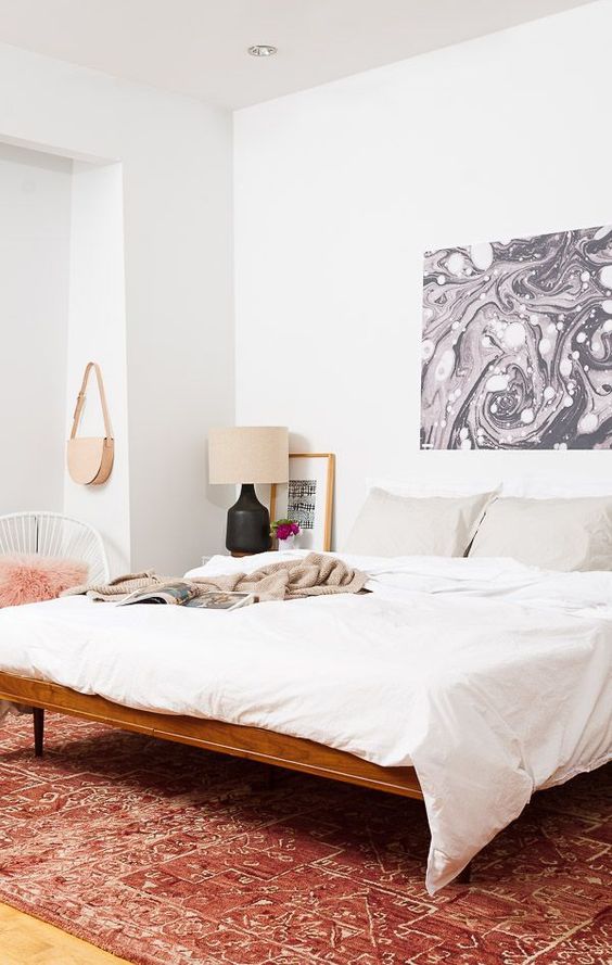 an eclectic bedroom with a bold printed rug, a stained bed with white bedding, a white chair, bold artwork and a lamp
