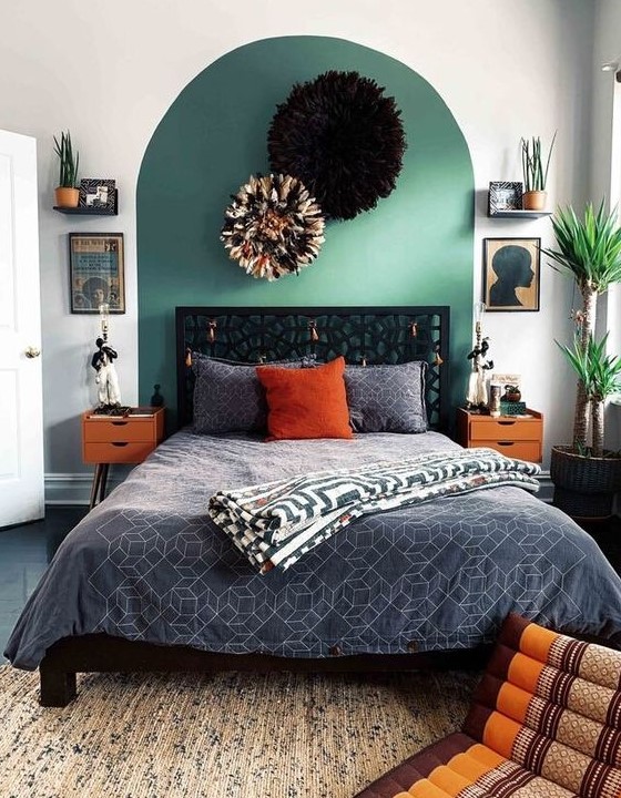 an eclectic bedroom with a green curved accent, a black bed with grey bedding, bold orange decor touches and nightstands, artwork, potted greenery and large pompoms
