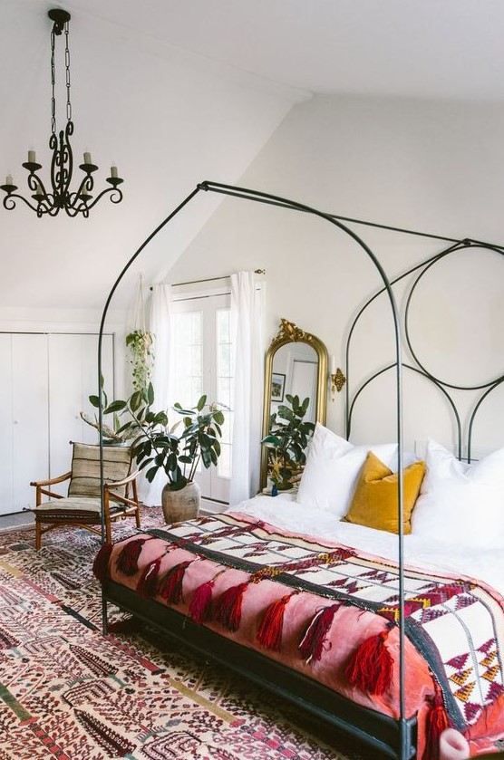 an eclectic bedroom with a large forged bed with colorful boho bedding, a bold boho rug, potted plant, a vintage chandelier on chain