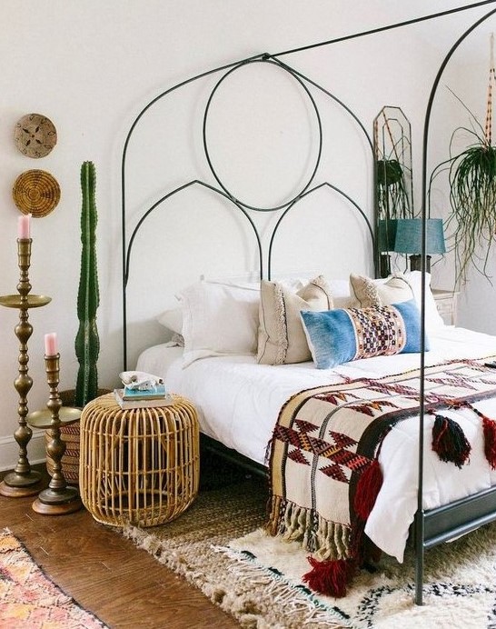an eclectic bedroom with a metal bed with decor, potted cacti and greenery, a rattan ottoman and vintage candle holders