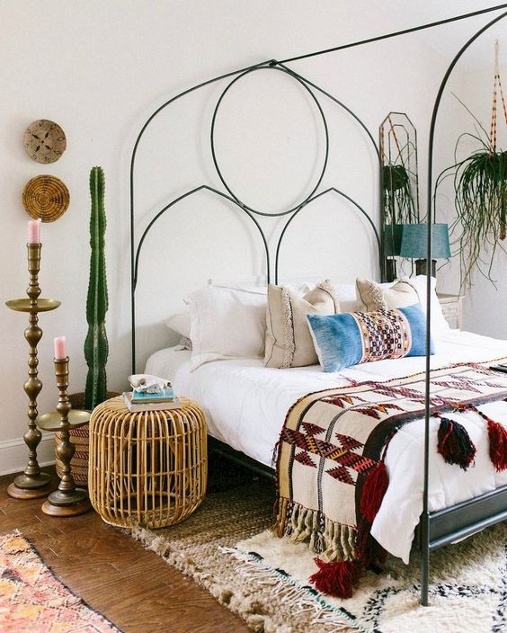 an eclectic bedroom with a metal bed with decor, potted cacti and greenery, a rattan ottoman and vintage candle holders