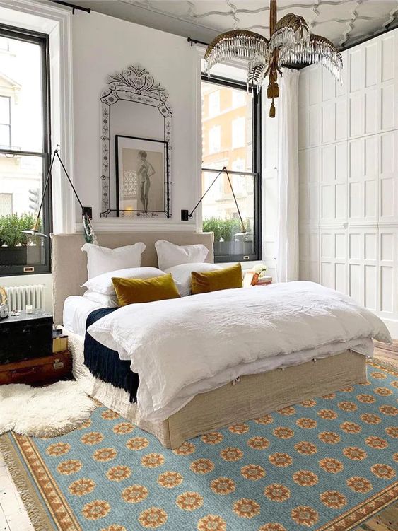 an eclectic bedroom with a paneled wardrobe, a neutral bed with neutral bedding, a bold printed rug, a chandelier and matching lamps