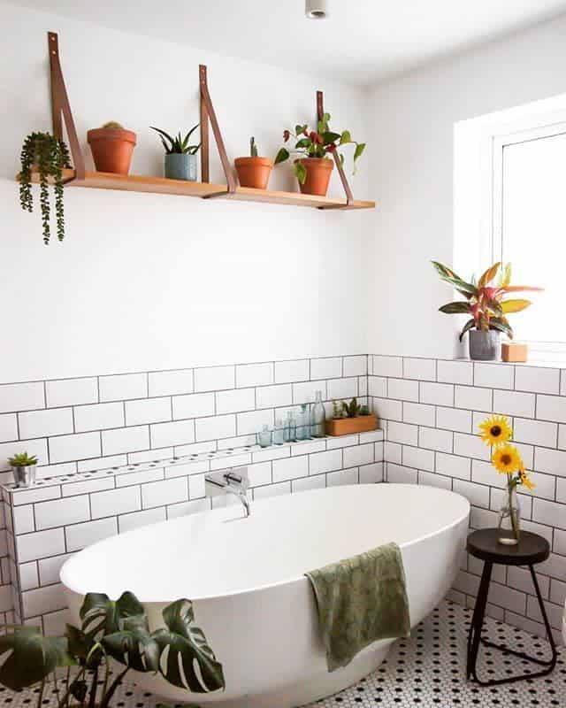 White tiled bathroom, freestanding bathtub, wooden wall shelf with potted plants 