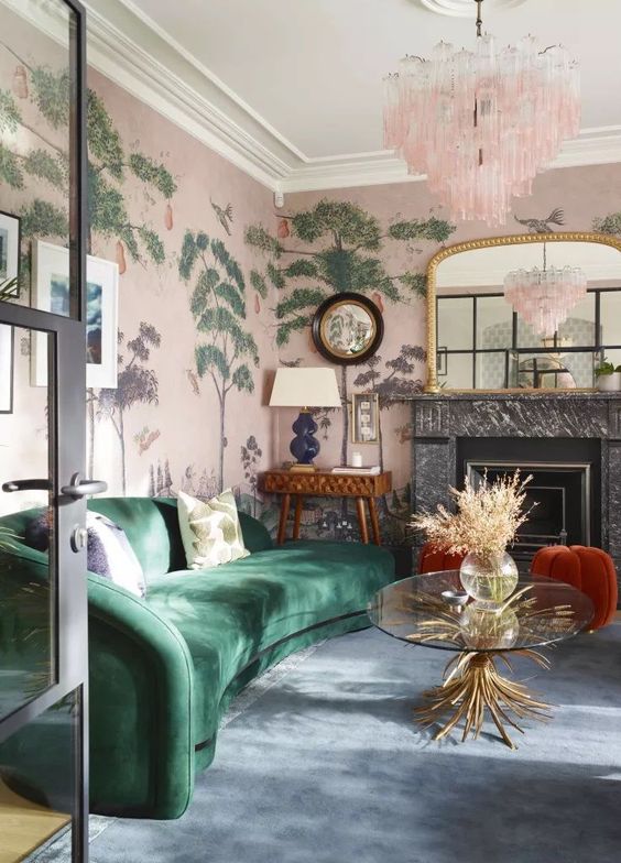 a whimsical living room with pink botanical wallpaper, a marble fireplace, a green curved sofa, rusty stools, a console table with a lamp and a pink chandelier
