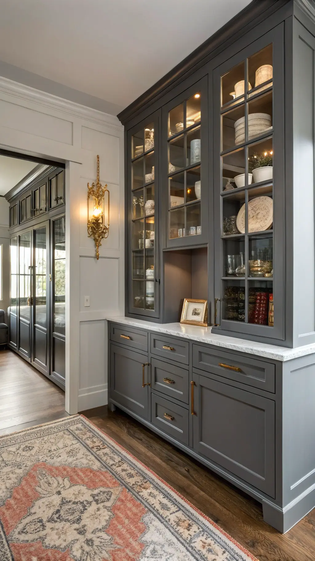 Butler pantry with floor-to-ceiling gray cabinets, white ironstone collection behind glass, brass library sconce, and vintage rug illuminated by afternoon light