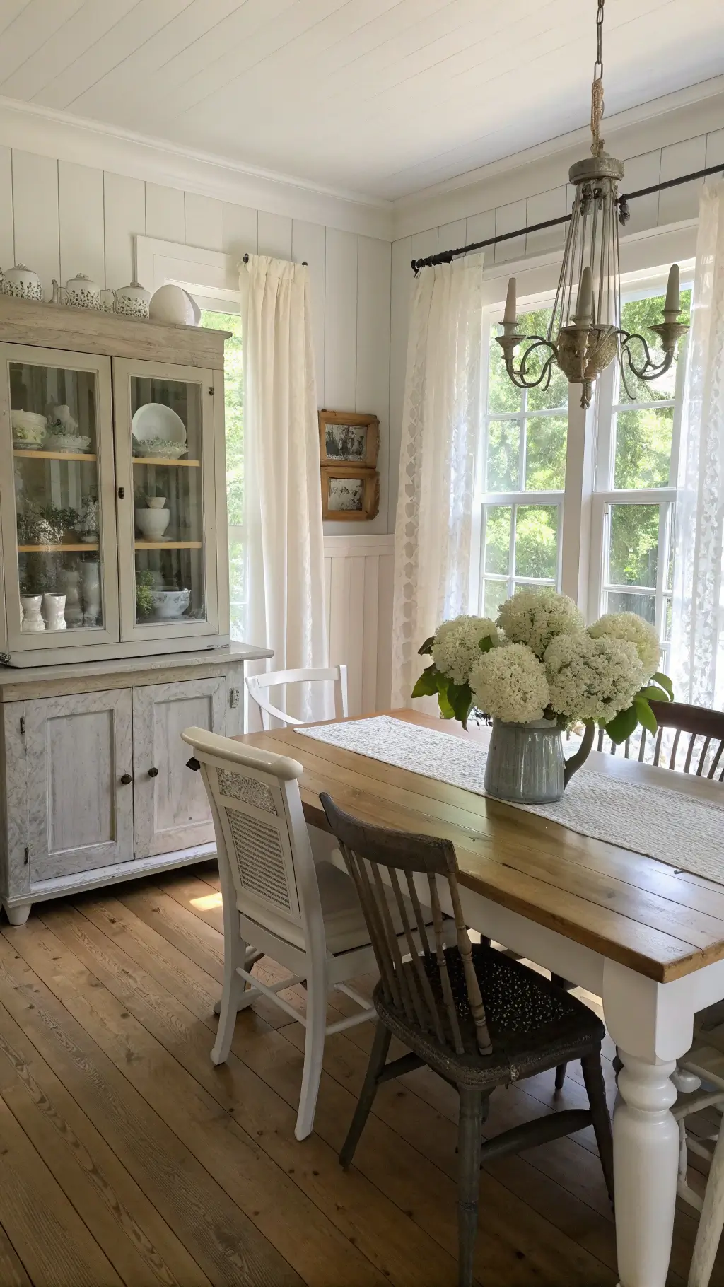Vintage-inspired dining room with creamy white walls, pine flooring, weathered oak farmhouse table seating eight, mixed chairs, antique hutch displaying ironstone collection, and dried hydrangeas centerpiece in a metal pitcher.