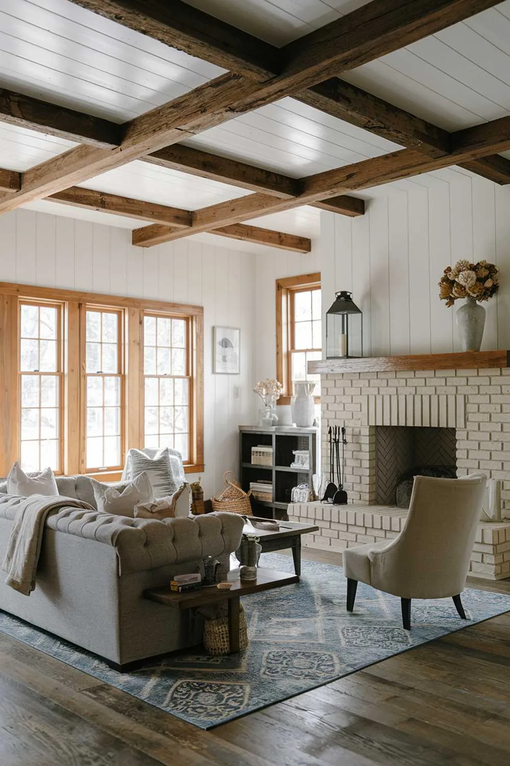 Modern farmhouse living room with rustic wooden beam ceiling, white walls, beige brick fireplace, gray couch, beige chair, wooden coffee table, and blue and white area rug.