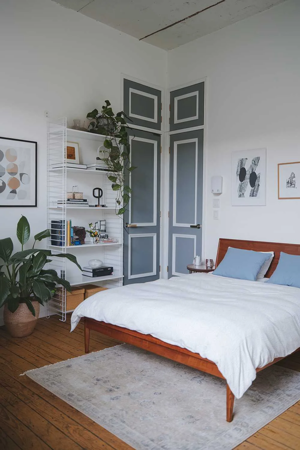 Modern Japandi bedroom with wooden floor, queen bed, white bookshelf filled with books and decor, large potted plant, beige rug, and artwork.
