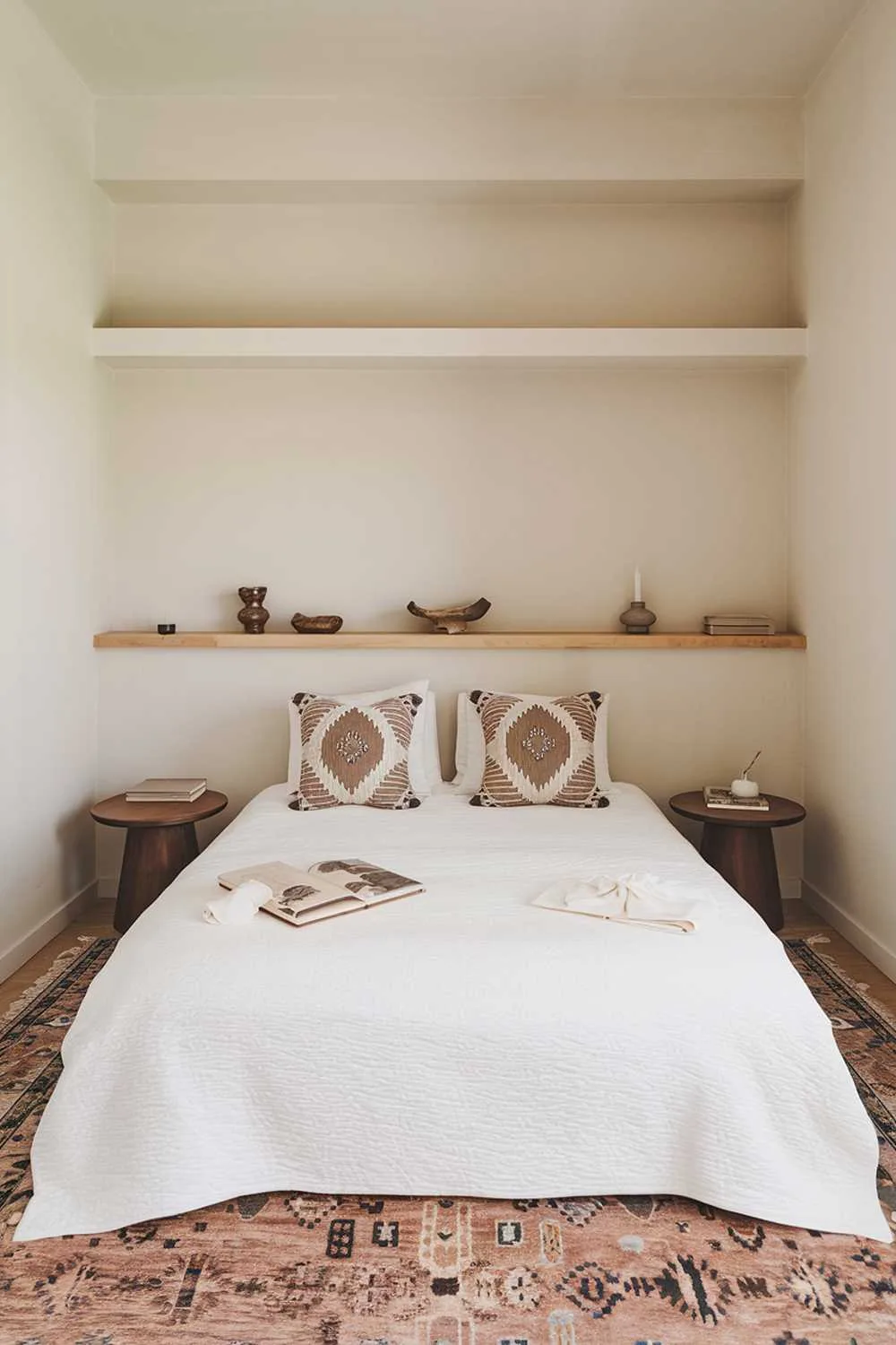 Small Japandi bedroom with queen bed, white duvet, two decorative pillows, matching wooden bedside tables, simple wooden headboard, beige walls, and large area rug.