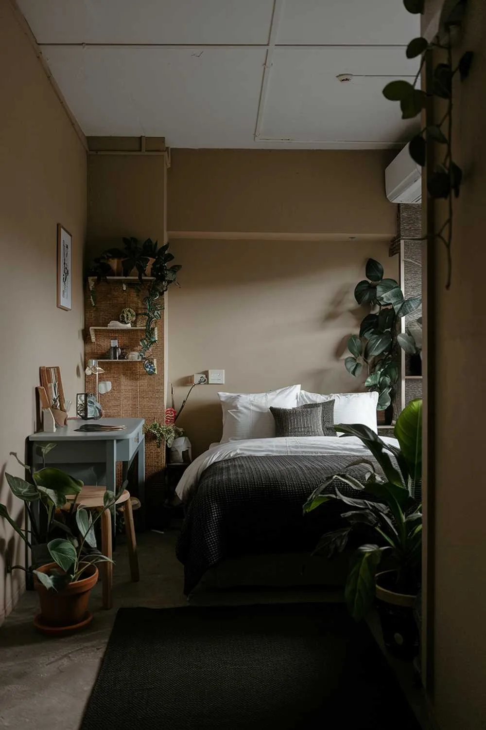 Small Japandi bedroom with low ceiling, queen bed with white sheets and dark duvet, small desk, medium-tone walls, dark rug, potted plants, and soft lighting.