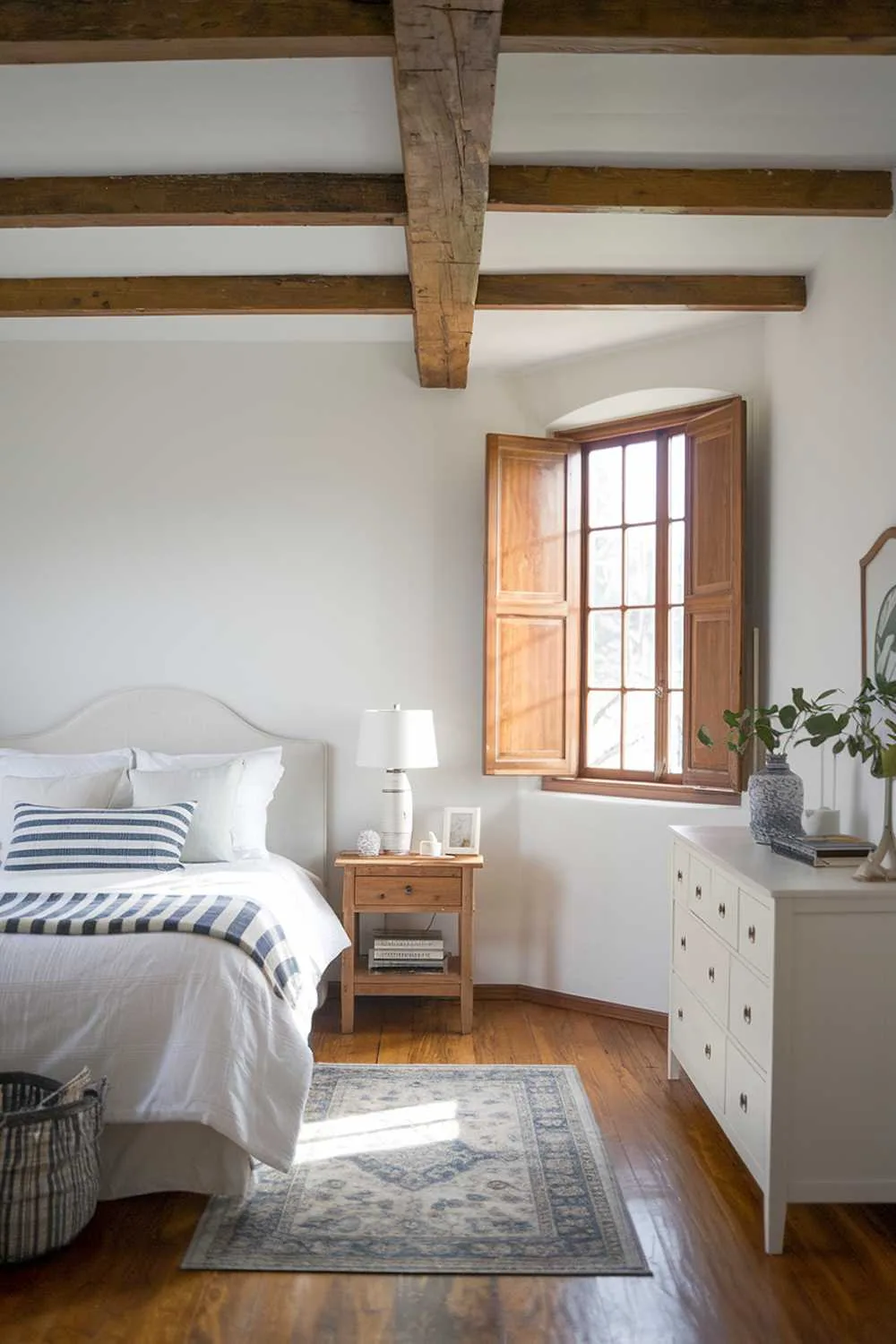 Scandinavian farmhouse bedroom with wooden beam ceiling, white walls, striped blue and white pillows, and wooden shutters.