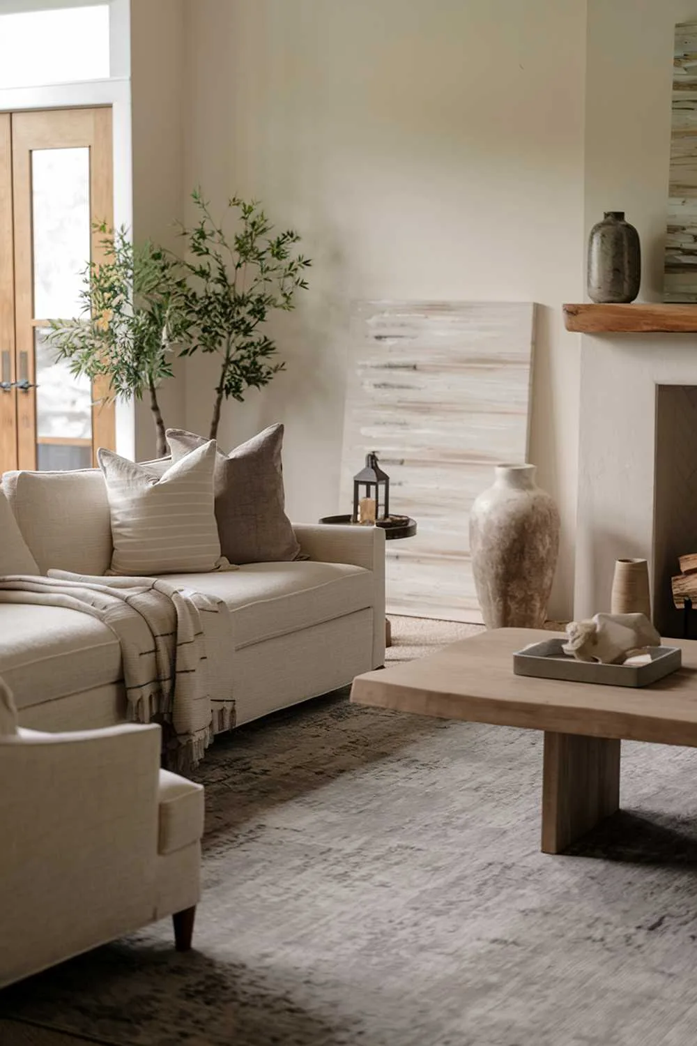 Neutral living room with beige sofa, assorted throw pillows, large gray rug, wooden coffee table, decorative art, potted plant, and fireplace.