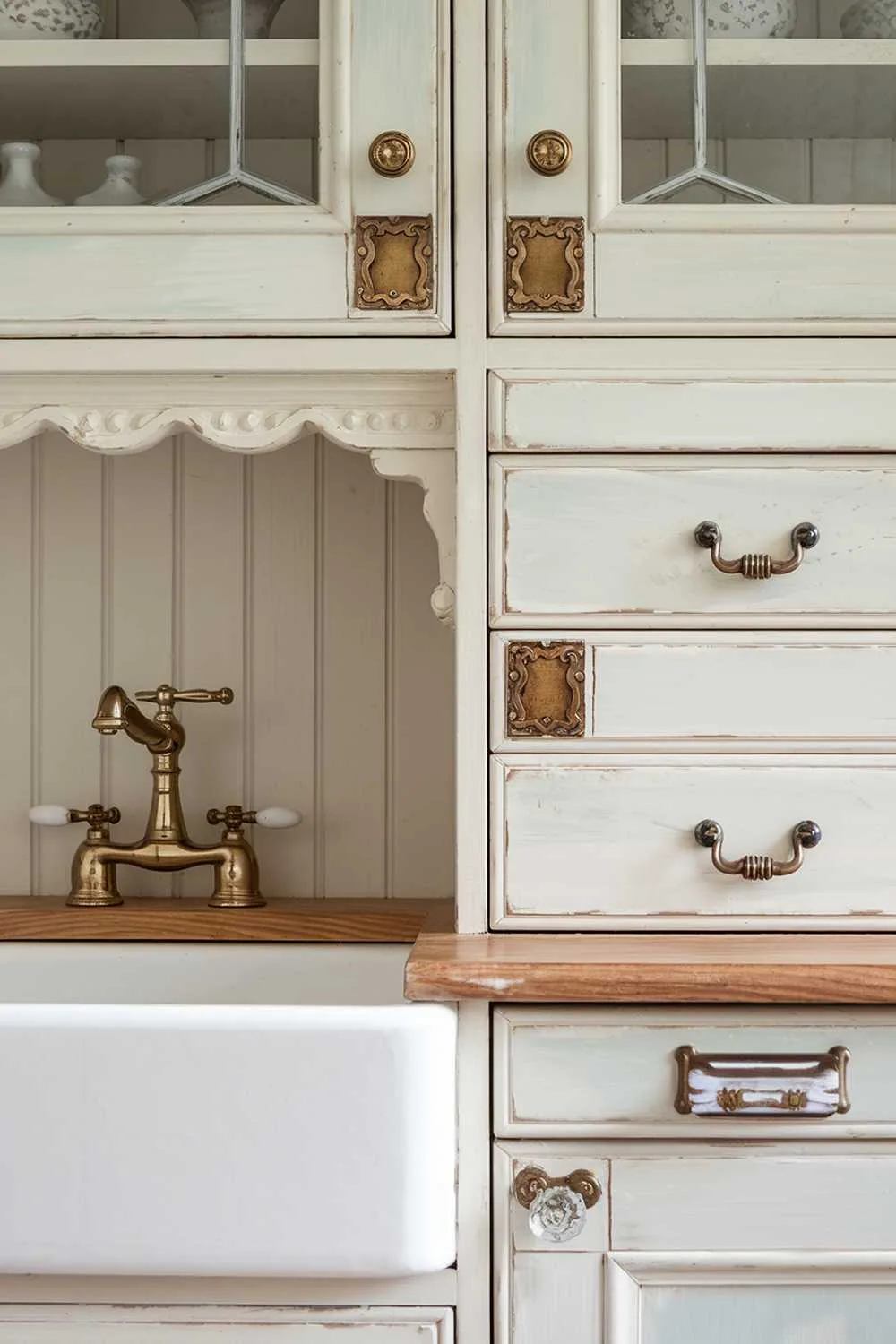 Cottage Kitchen Hardware Details Close-up of pale blue cabinet doors with vintage brass bin pulls and glass knobs, decorative hinges, a Victorian-style aged brass faucet over a white farmhouse sink, beadboard backing, and delicate trim work.