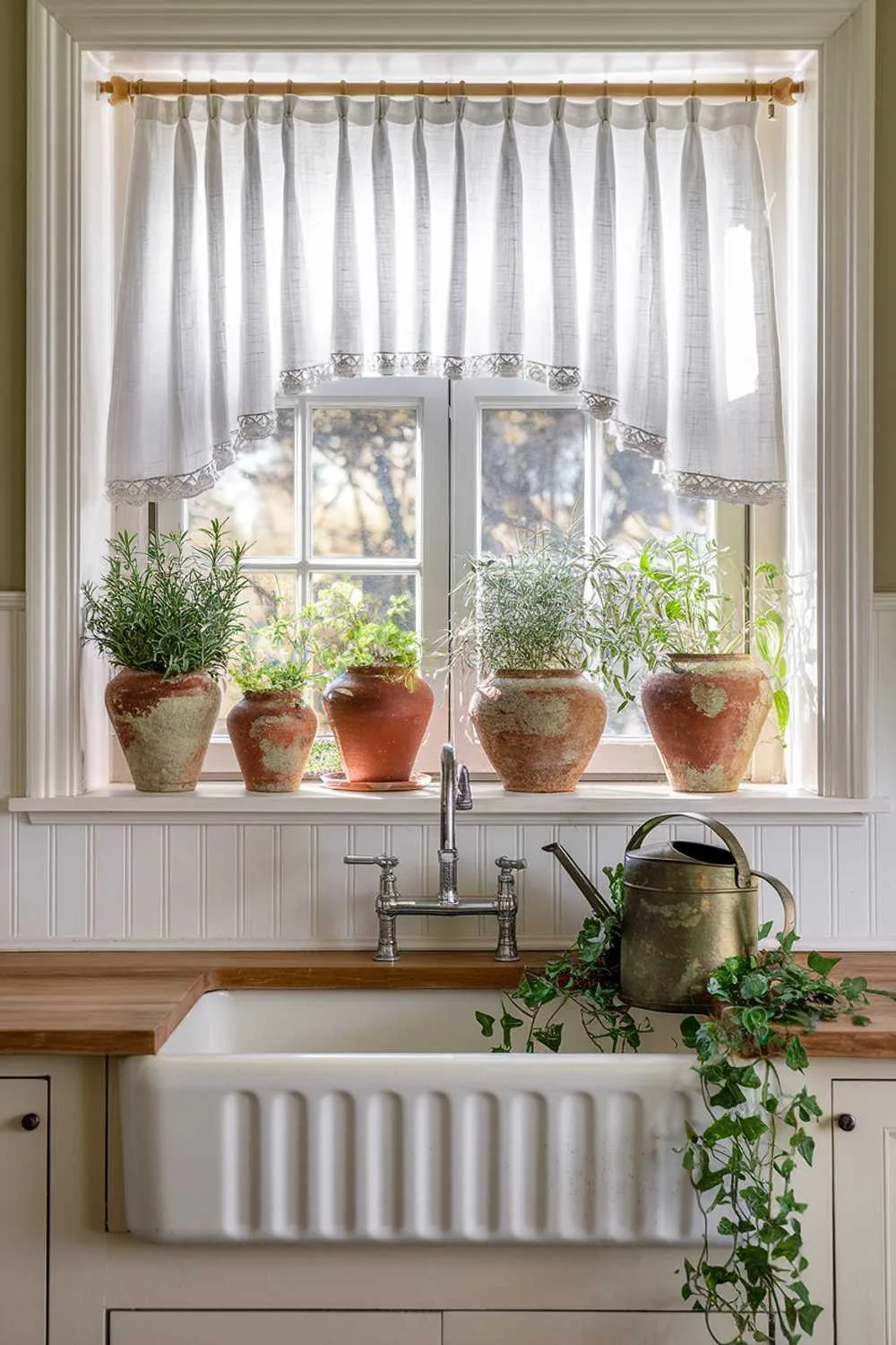 Window Treatments for Cottage Kitchens Sunlit kitchen window with white linen cafe curtains filtering light onto a farmhouse sink, a deep windowsill holding terracotta pots with fresh herbs and a vintage watering can, white beadboard walls, and a vintage-style aged brass faucet.