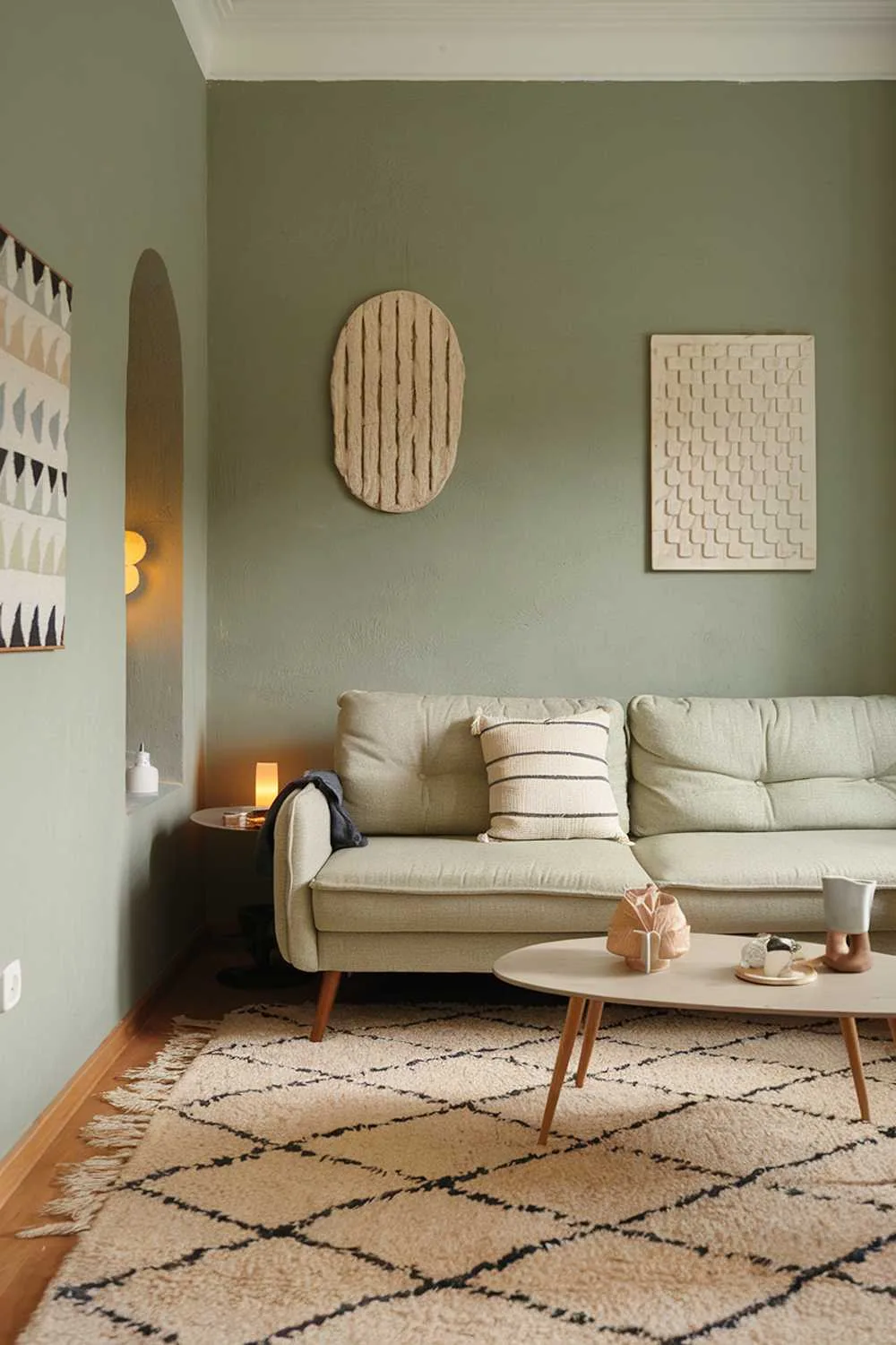 Living room with sage green walls, beige sofa with cushions, geometric beige rug, white coffee table, warm lighting, and decorative art pieces.