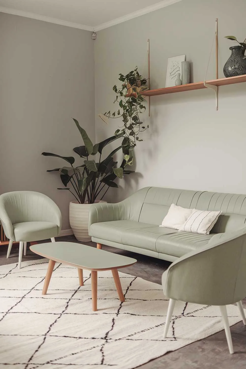 Sage green sofa with white cushions, wooden coffee table with white top, two sage green chairs with white legs, wooden shelf with decor, large potted plant in white pot, white rug, light gray walls, soft lighting.