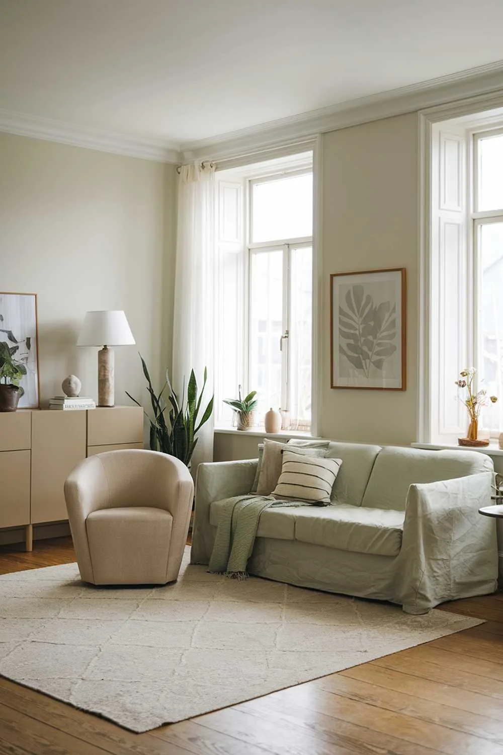Cozy living room with sage green sofa, beige area rug, beige sofa with throw pillows, beige chair, beige cabinet, beige lamp, green plant, white walls and ceiling, large windows with white curtains, wooden floor.