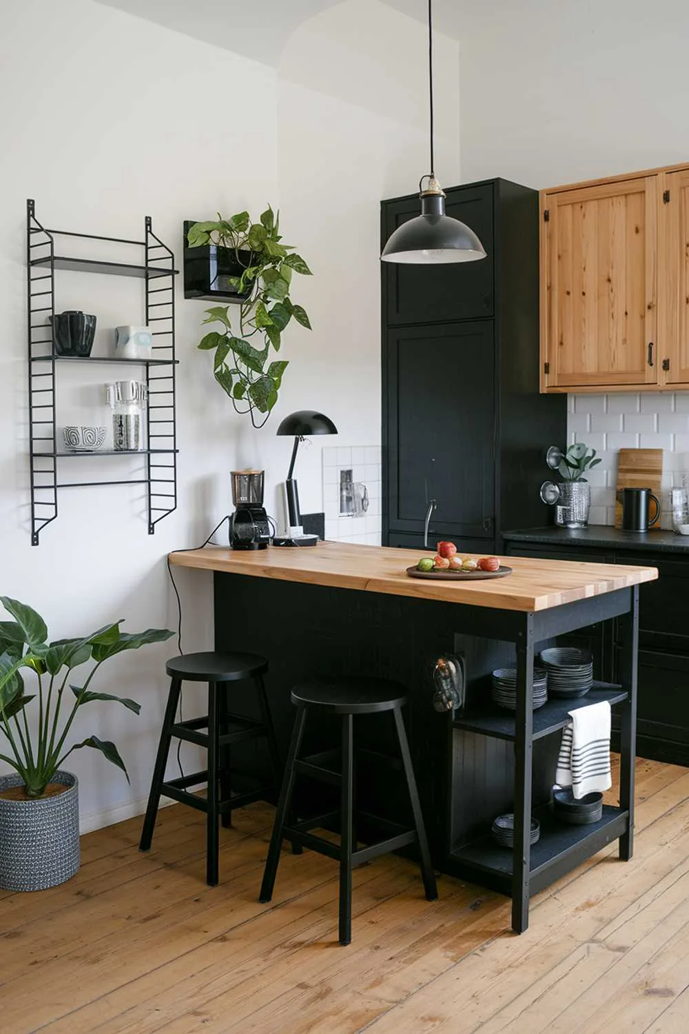 Black kitchen island with natural wood top, black stools, black pendant light, and potted plant on wall shelf