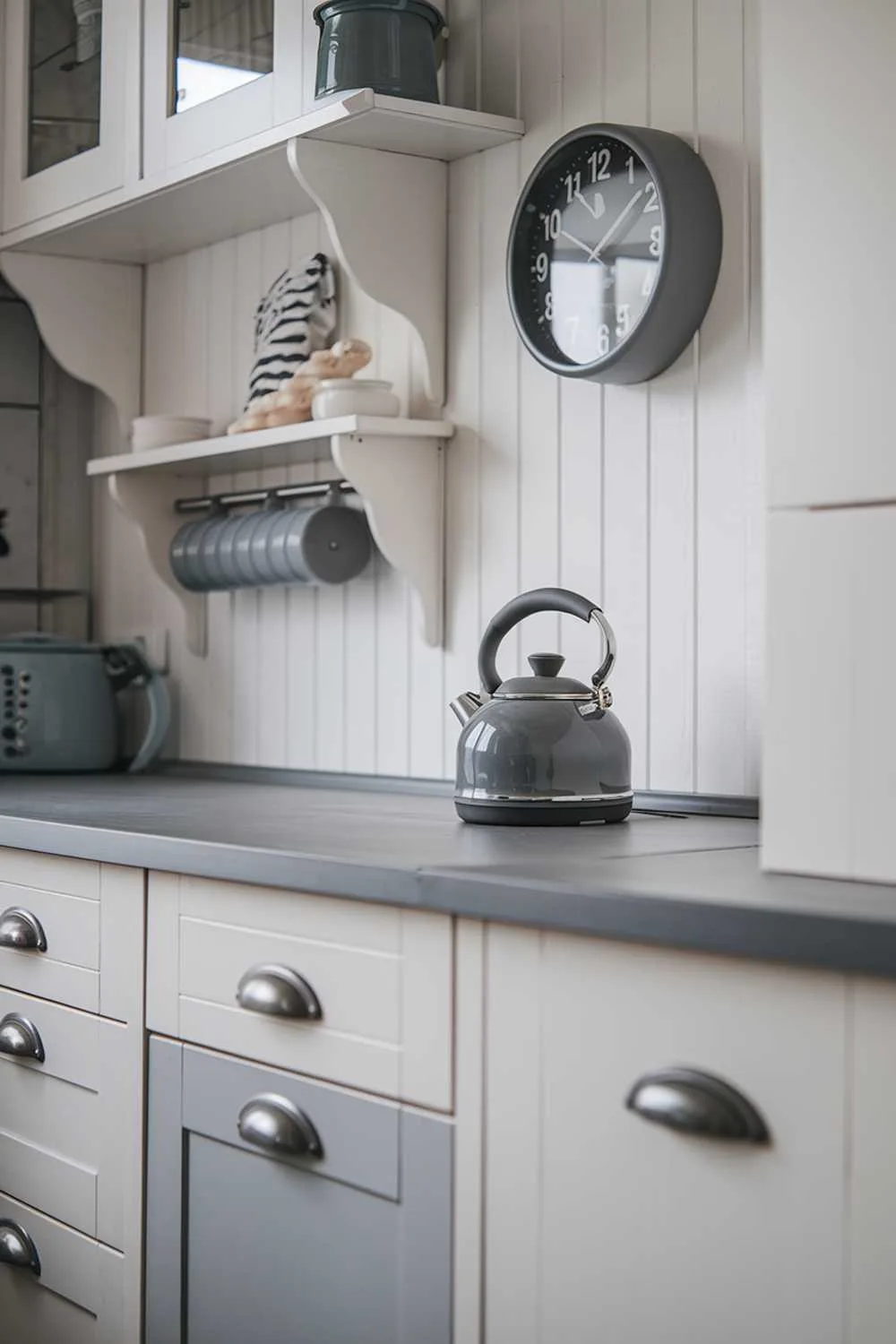 Close-up of white cabinets with gray drawers, gray countertop, gray kettle, and wall shelves.