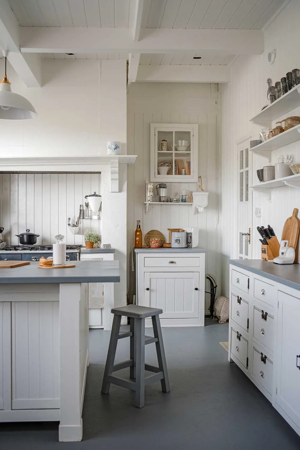 White kitchen with gray countertop island, gray stool, white cabinets, and pendant lighting.