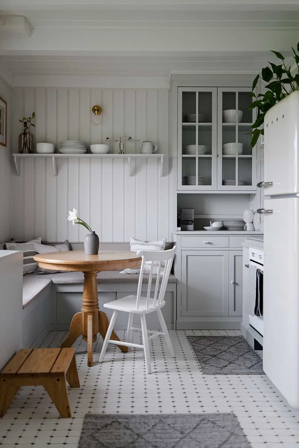 Breakfast nook with round wooden table, white chairs, gray bench, glass-front cabinets, and potted plants.