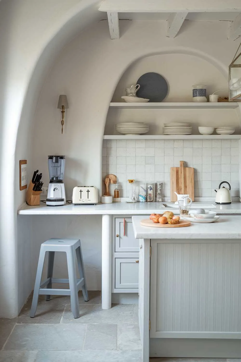 White kitchen with smoothie maker, toaster, coffee maker on countertop, gray stool, and light gray island.