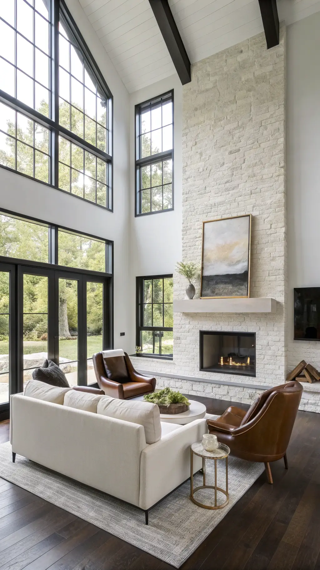 Modern farmhouse living room with double-height windows, cloud-white walls, and dark hardwood floors, featuring contemporary leather chairs, oversized linen sofa and abstract canvas above a limestone fireplace; accented by geometric pottery and brass.