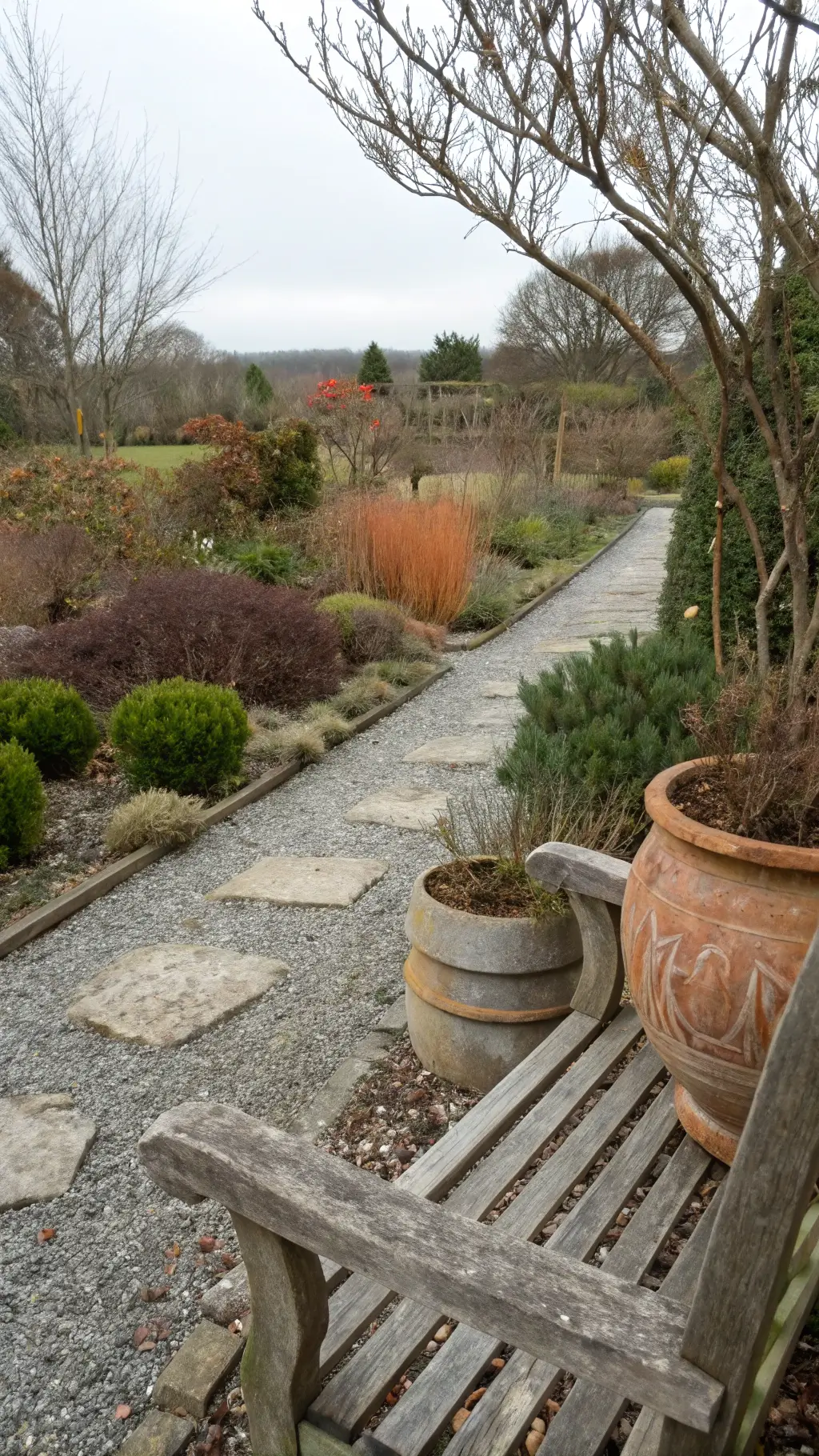 Midday overcast view of a gravel garden with soft rake patterns, framed by native shrubs showing seasonal die-back, featuring a weathered wooden bench and hand-thrown pottery nestled in groundcover.