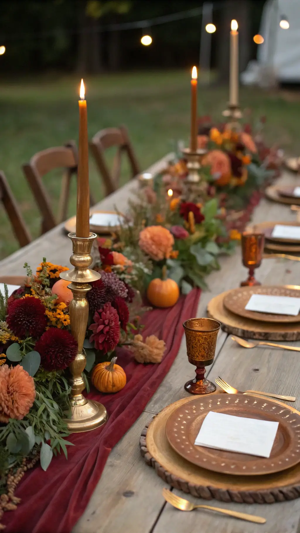 Intimate harvest table with burgundy dahlias, orange ranunculus, mini pumpkins, vintage brass candlesticks, and rust napkins under warm Edison bulb lighting