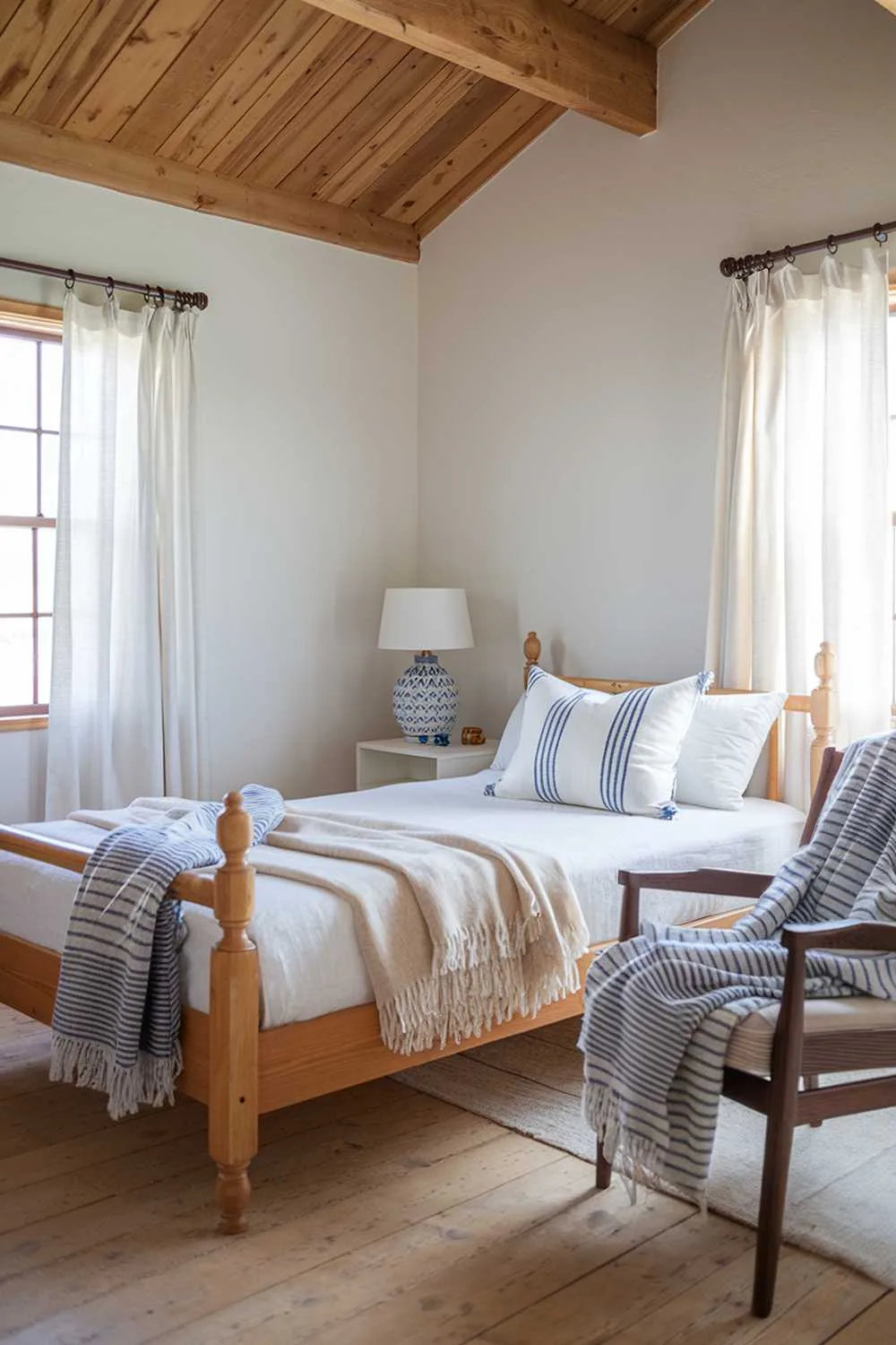 Scandinavian farmhouse bedroom with exposed wooden beams, white walls, and wooden floors. Wooden bed frame with white bedding and blue-striped pillows.